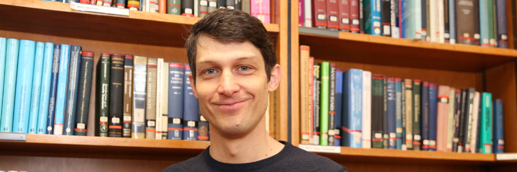 Professor Ryan Cook looking straight to camera and smiling. He is stood in front of shelves of books.