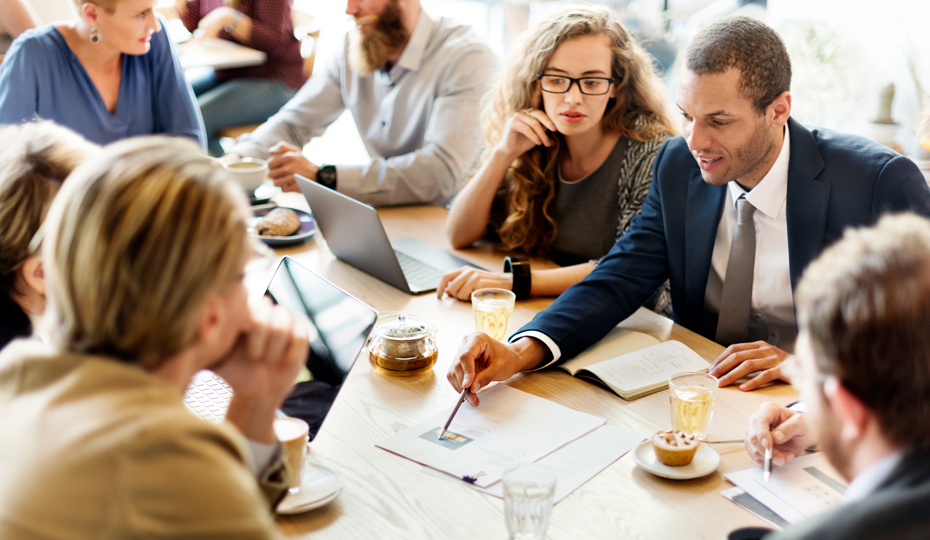 A business team meeting with team members collaborating around a busy table strewn with papers and handouts