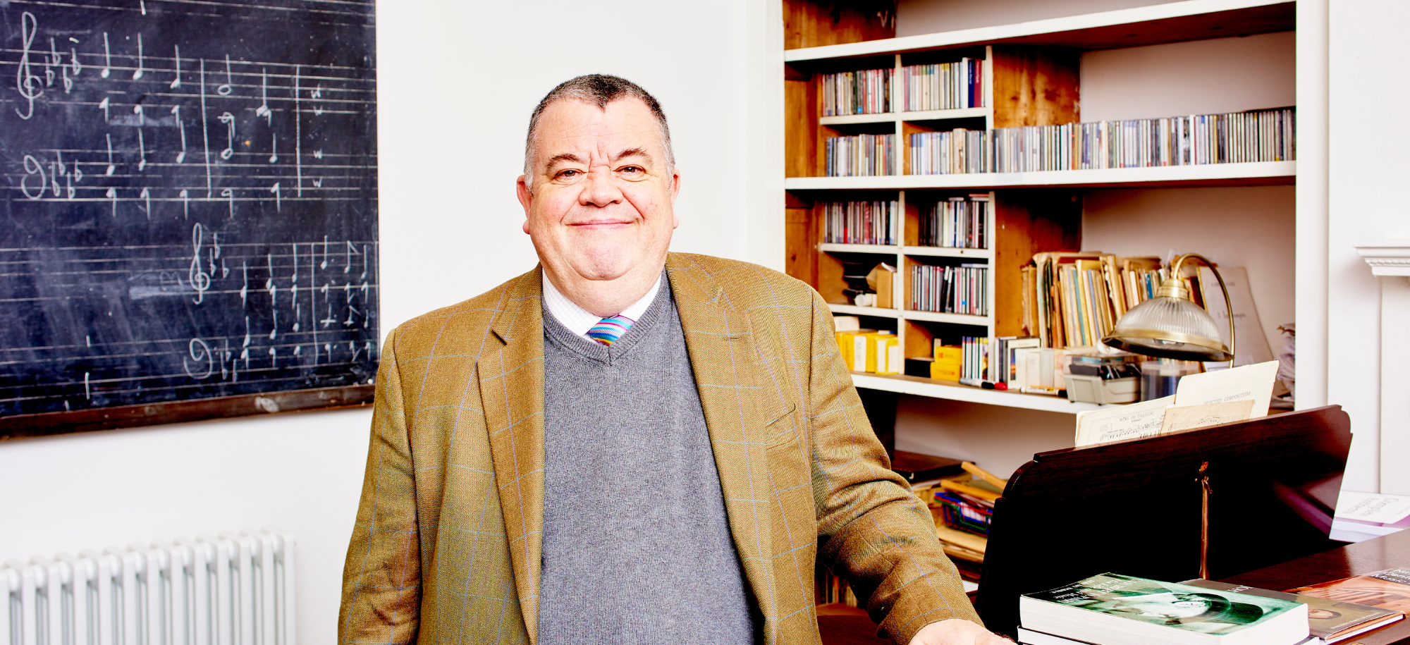 Professor Jeremy Dibble standing in front of a bookcase and a blackboard with musical notes written on it