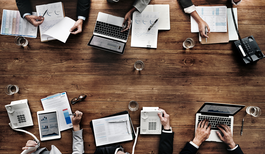 A meeting with laptops, circulars and telephones scattered around a busy table