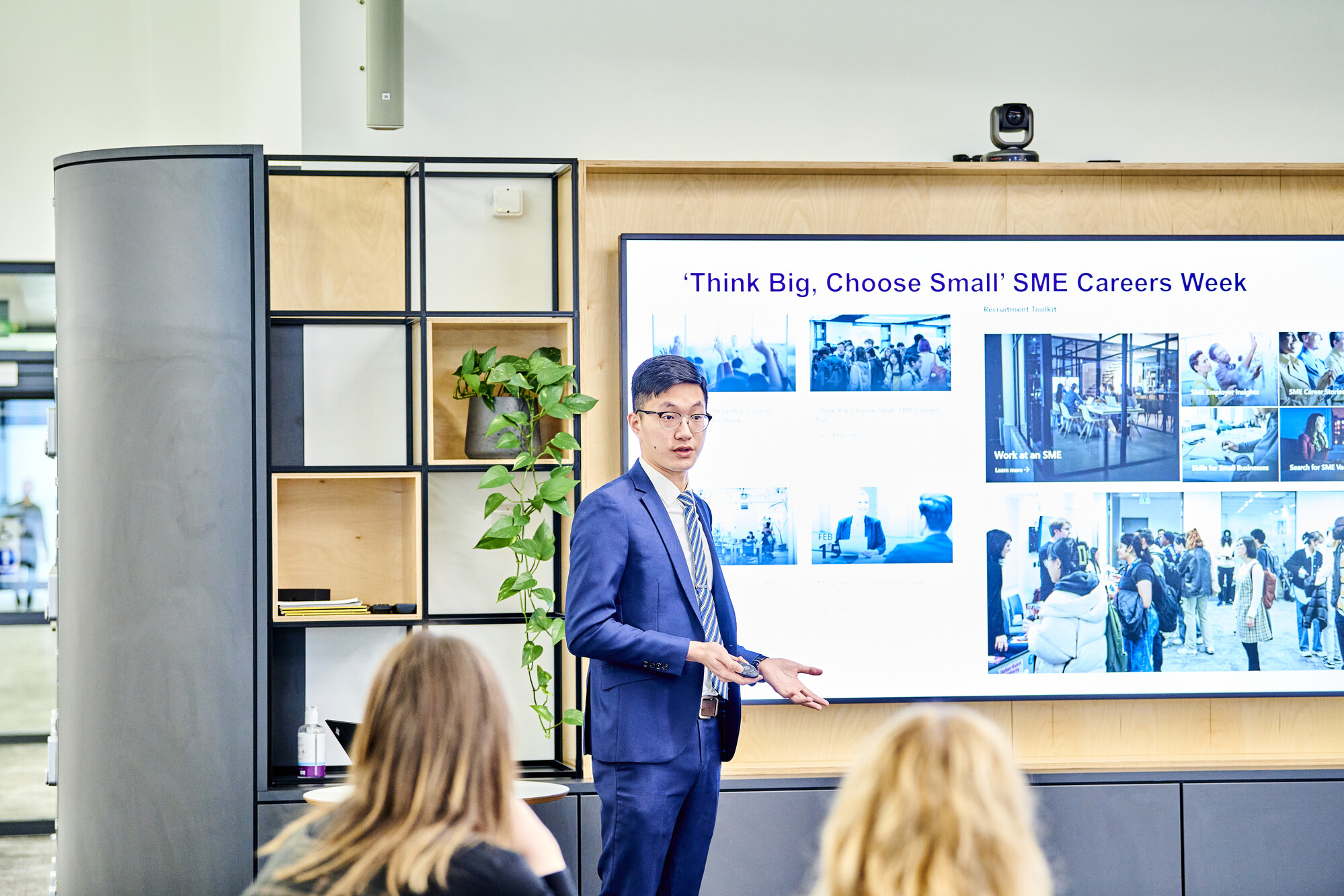 Young man in a business suit stands in front of a digital screen. The screen shows a collage of images and the words 