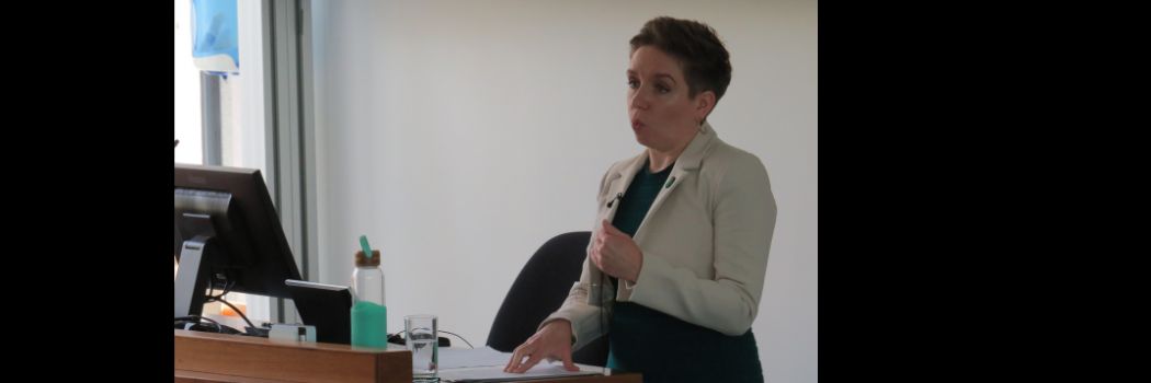 Woman standing at presentation desk, addressing audience (off camera)