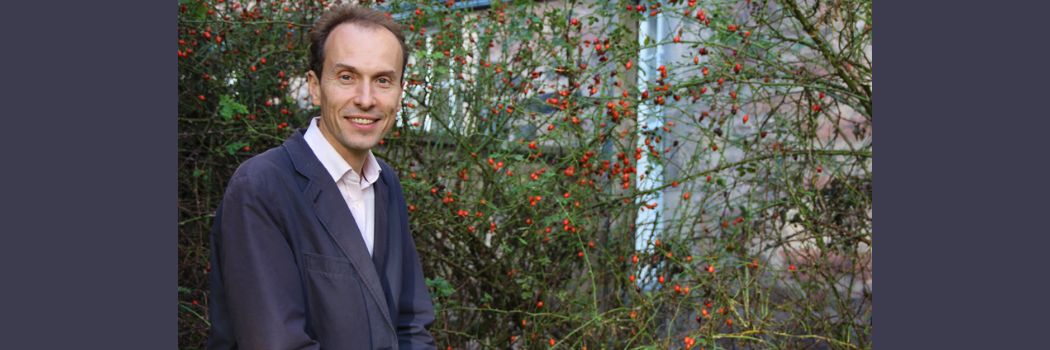 Professor Jolyon Mitchell, wearing a white shirt and dark blue jacket, smiles at the camera standing in front of a green shrub with red berries.