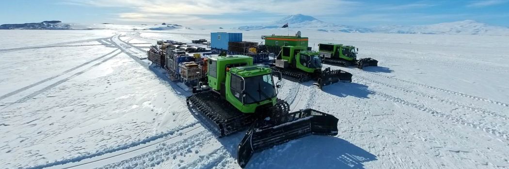 Green big track snow ploughs stand on the snow and ice sheet with a white and blue sky behind them.