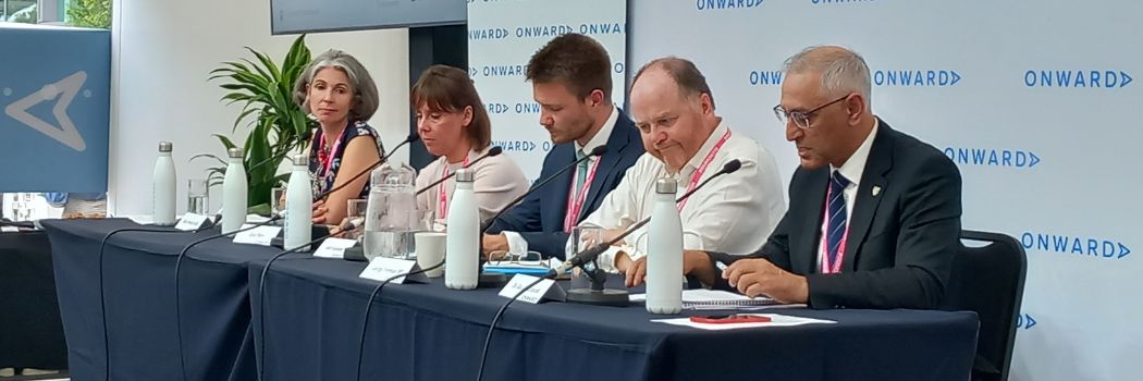 Five people sitting behind a table, with the man on the front right addressing an off-picture audience