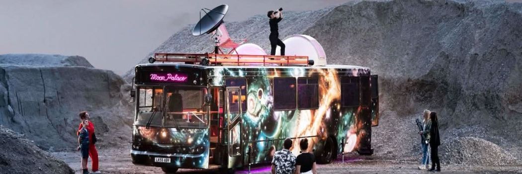 A single decker bus spray painted with images of space and featuring a satellite dish on its roof is parked in a dusty, moon-like landscape.