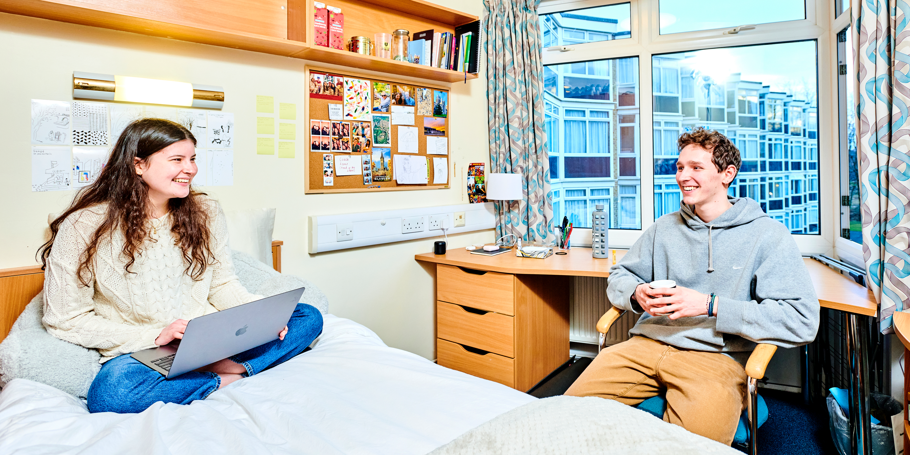 Two students sit in a typical college bedroom. She sits crosslegged on her bed, holding a laptop. He sits on a chair next to the bed and they are talking and laughing