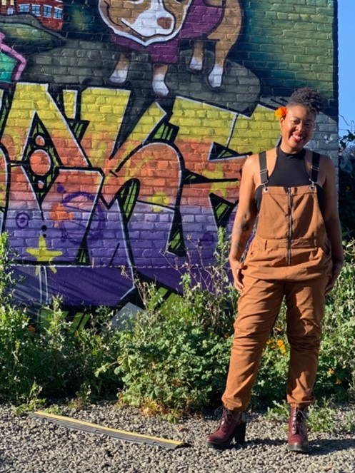 A smiling Black person stands in warm sunlight wearing a black top and rust orange dungarees with reddish brown heeled boots. They have their hair gathered up and red and orange flowers above their right ear. Behind them is a wall with colourful graffiti and green bushes.