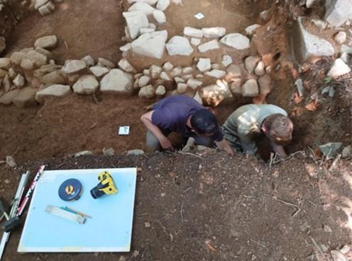 TTwo people crouched in an archaeological trench, drawing a section of deposits. Behind them are the remains of a building. On the edge of the trench is some drawing equipment.