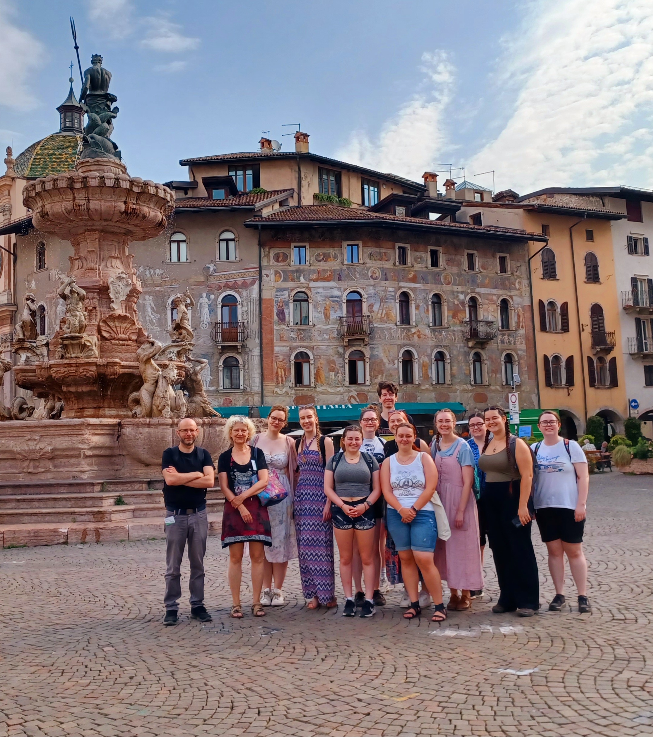 Group photo of 13 smiling people in summer outfits, standing in a piazza in Trento, Italy, adjacent to a large fountain of the Roman god Neptune.