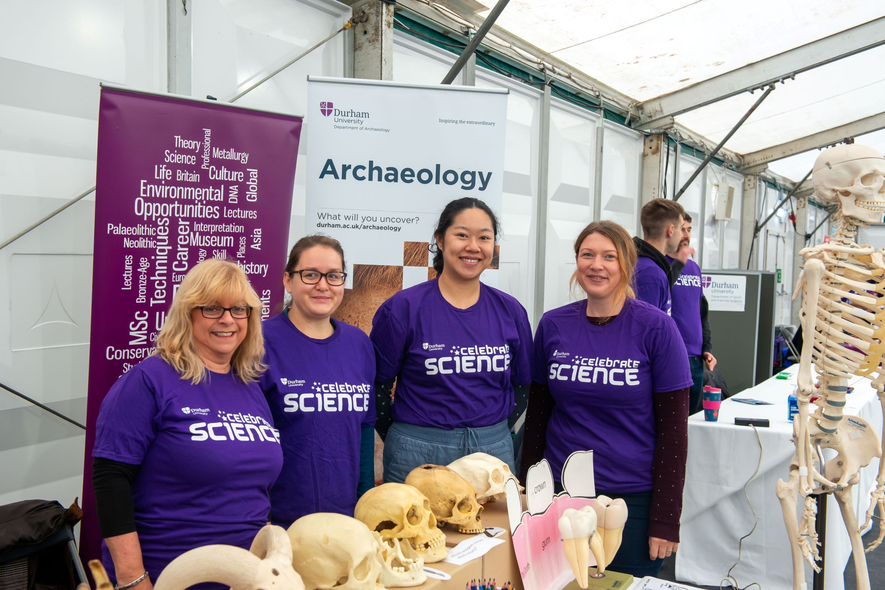 Group photo of four smiling Archaeology volunteers in purple shirts, standing behind a table with animal and human skull models on display. Behind them are pull-up banners advertising the Department of Archaeology.