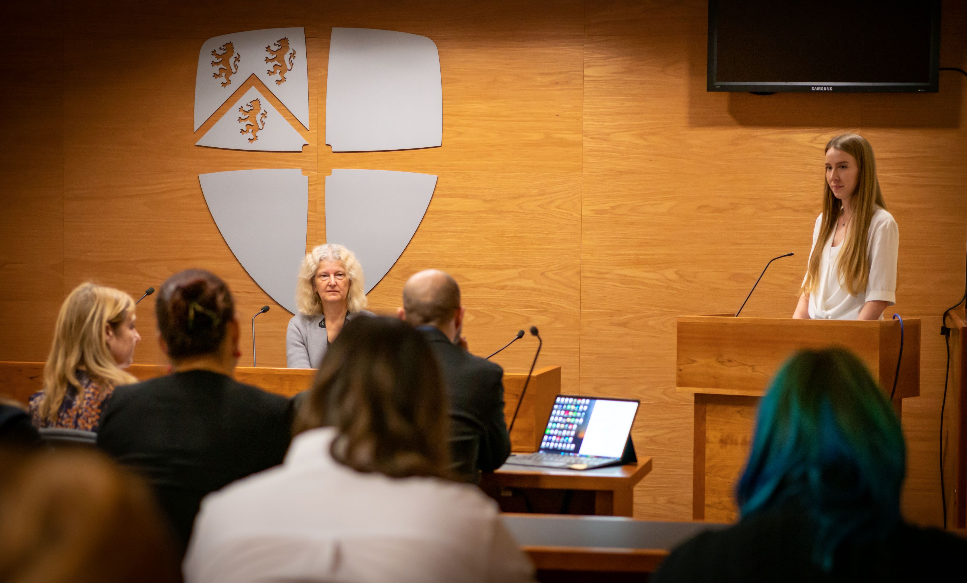 In a mock court room, a student stands at a podium delivering an Expert Witness Statement. Other staff and students are sat behind benches.