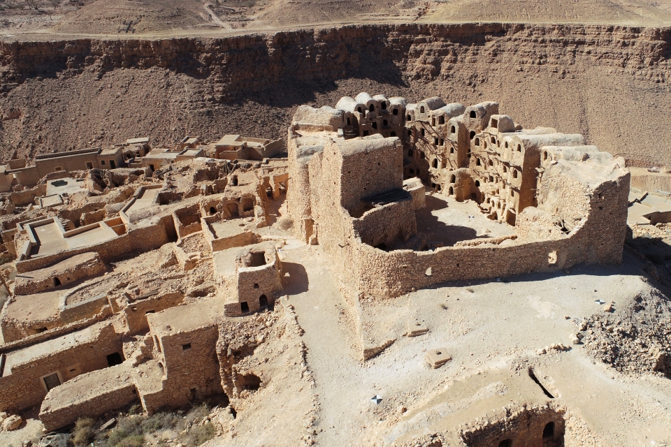Under strong golden sunlight are archaeological remains of buildings made of stone, gypsum, and red mud. A cliff and the base of mountains are visible in the distance.