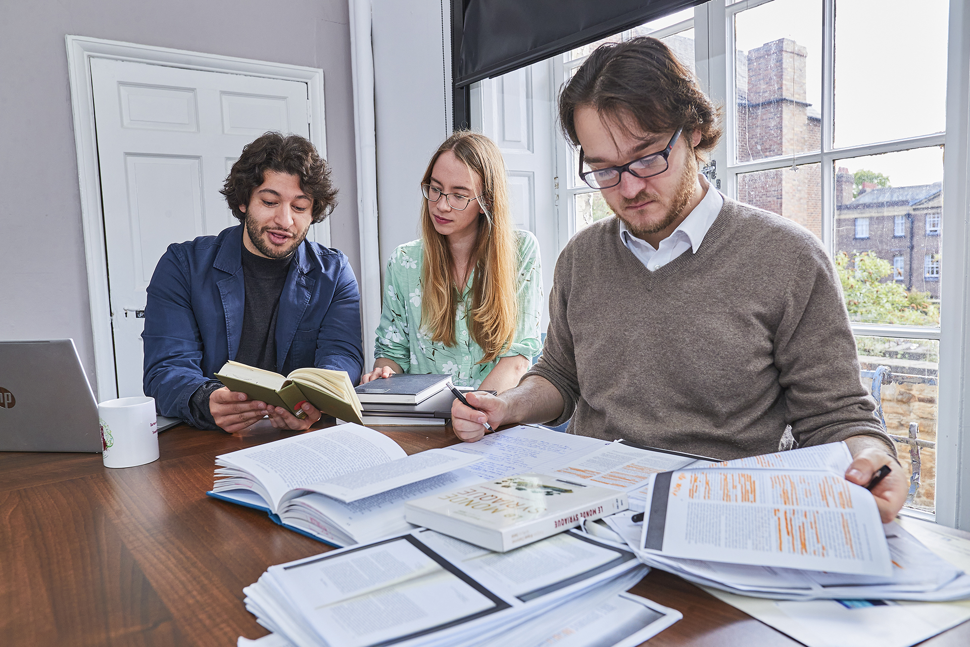 Three PhD students, two men and a woman, sit at a table with an open laptop and books and papers spread around. In the foreground on the right, a student looks at a printed article with highlighting, while in the background the other two are reading a book