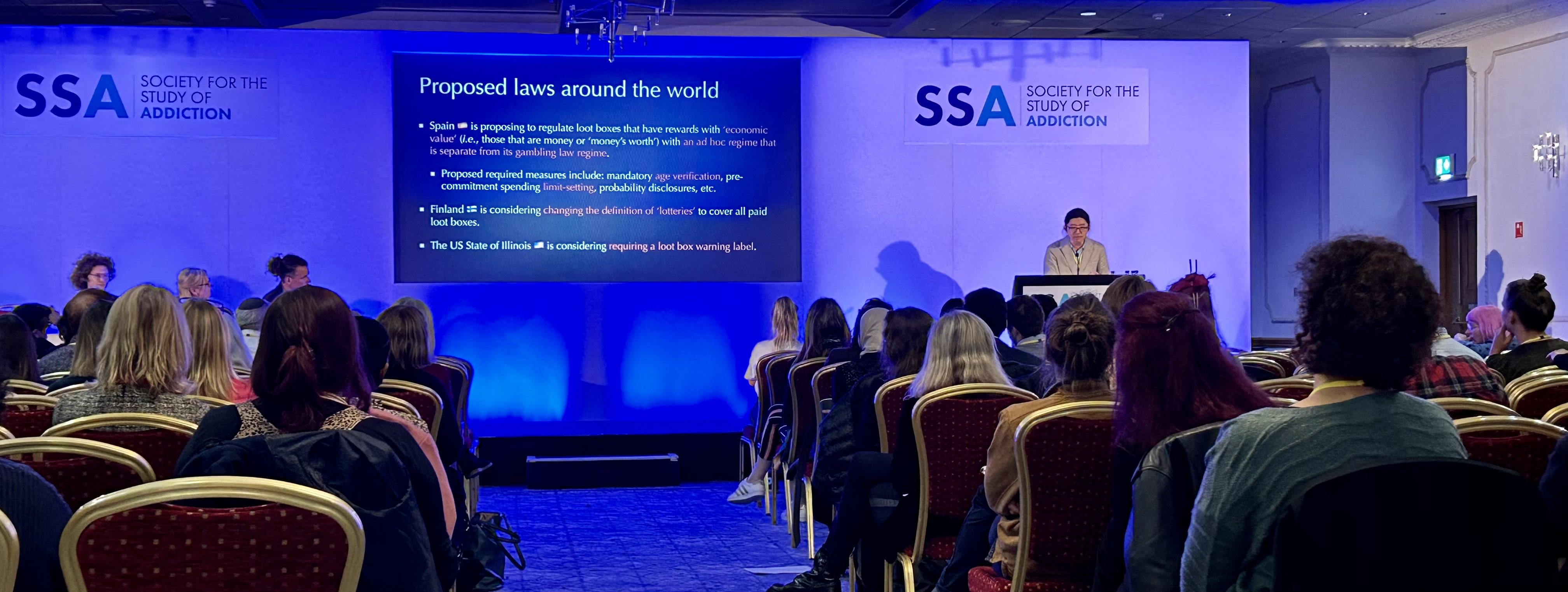 Leon speaking at a conference. The room is lit in blue and Leon is wearing a beige suit with his long dark hair tied back.