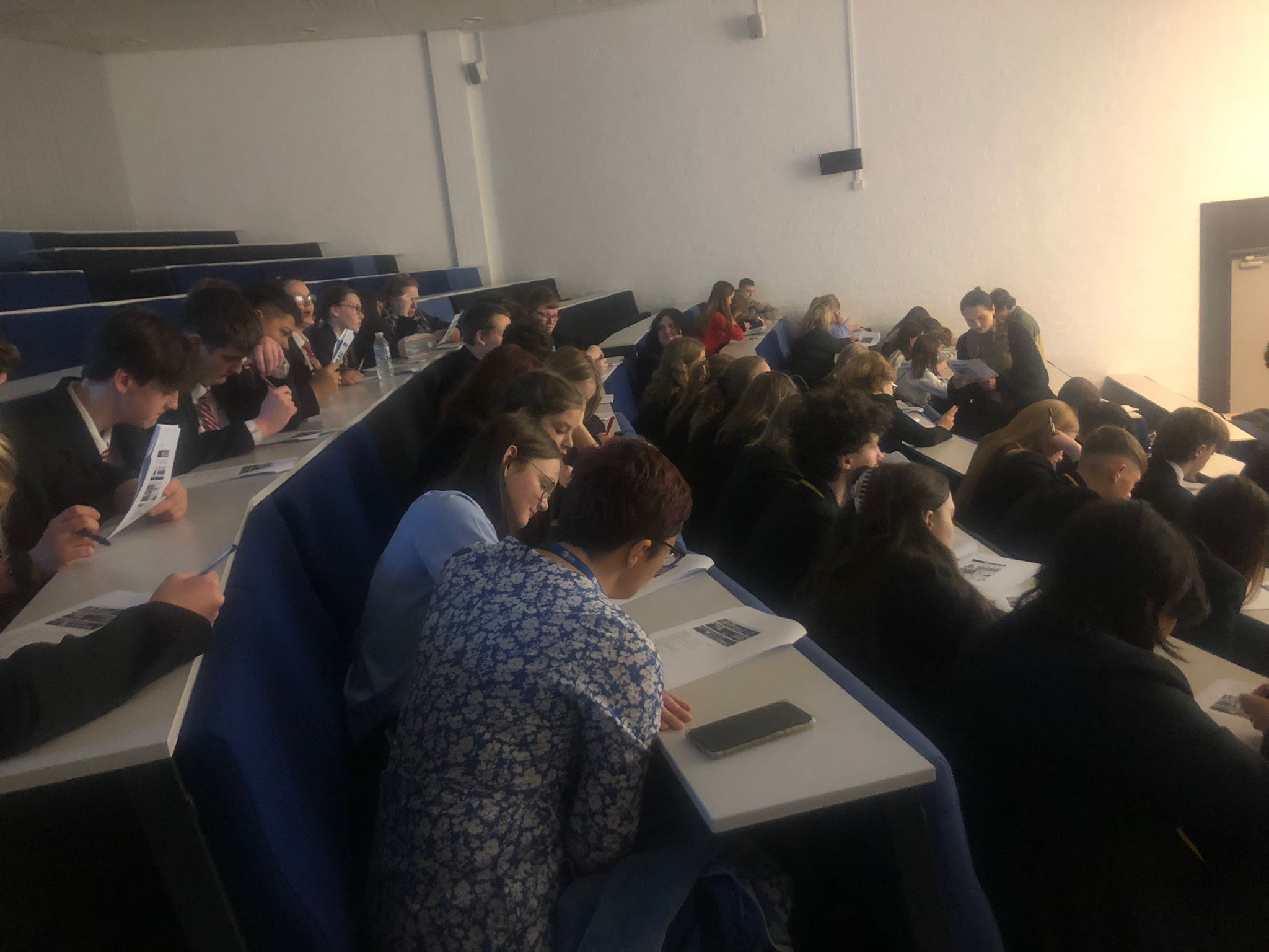 School pupils sitting in rows in tiered university lecture theatre writing.