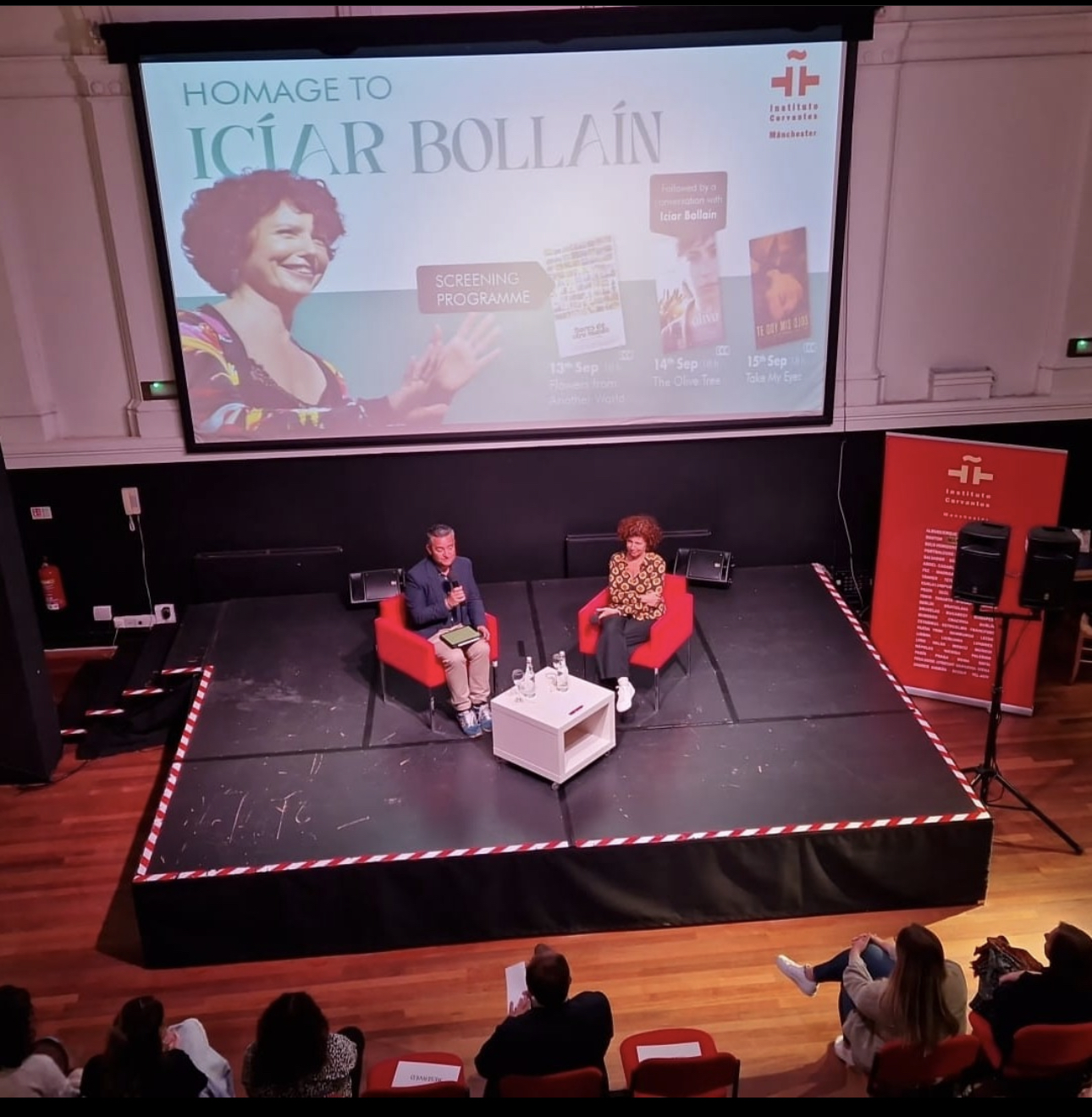 Two people sitting on a stage with a large screen behind them