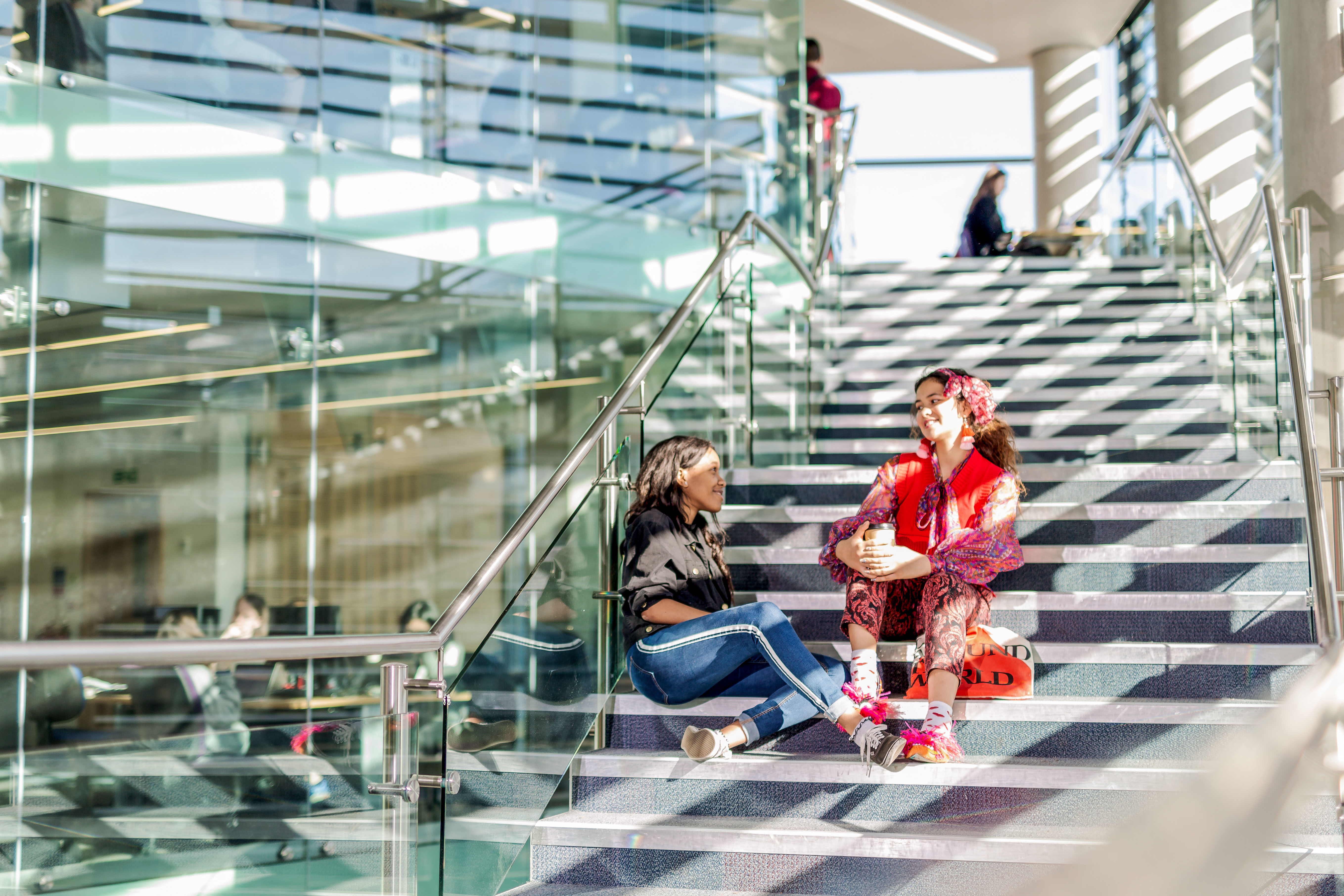 Two female students sit on sunlit library stairs chatting