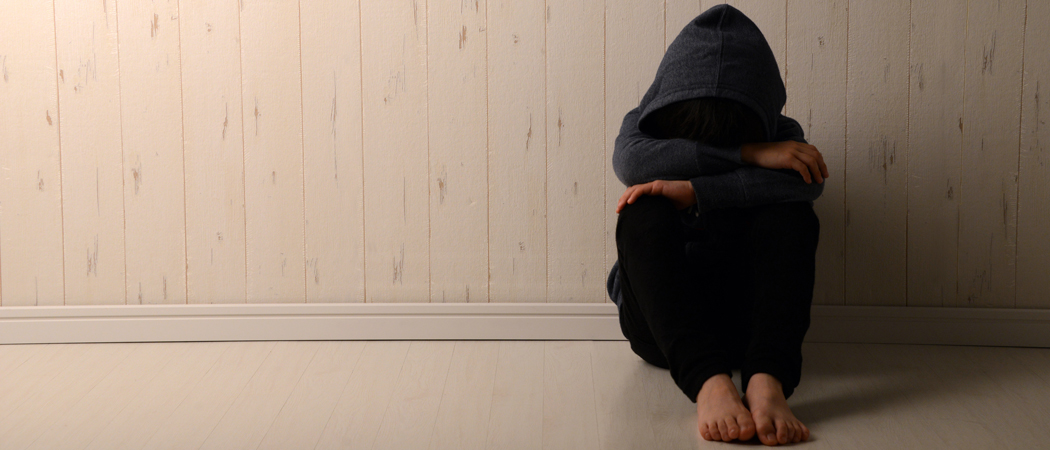 An upset child sitting against a wall with a hood pulled tightly over his head