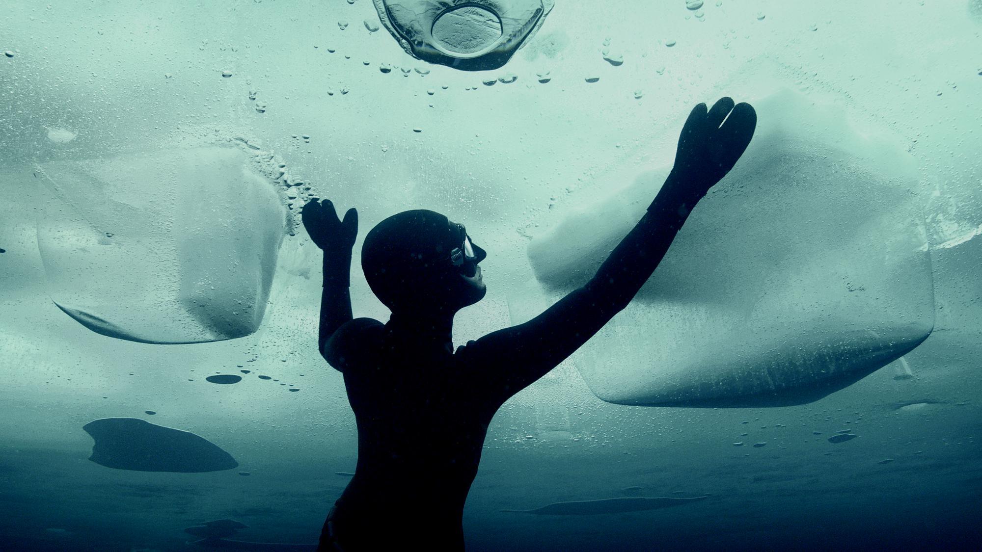 A man in a diving suit and goggles reaches up through water to an ice sheet over his head