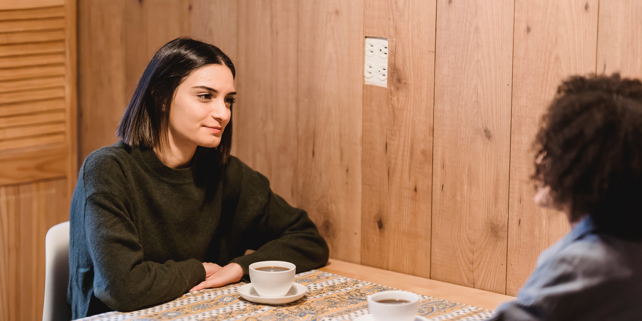 Two people sitting at a table talking to each other, with cups in front of them on the table