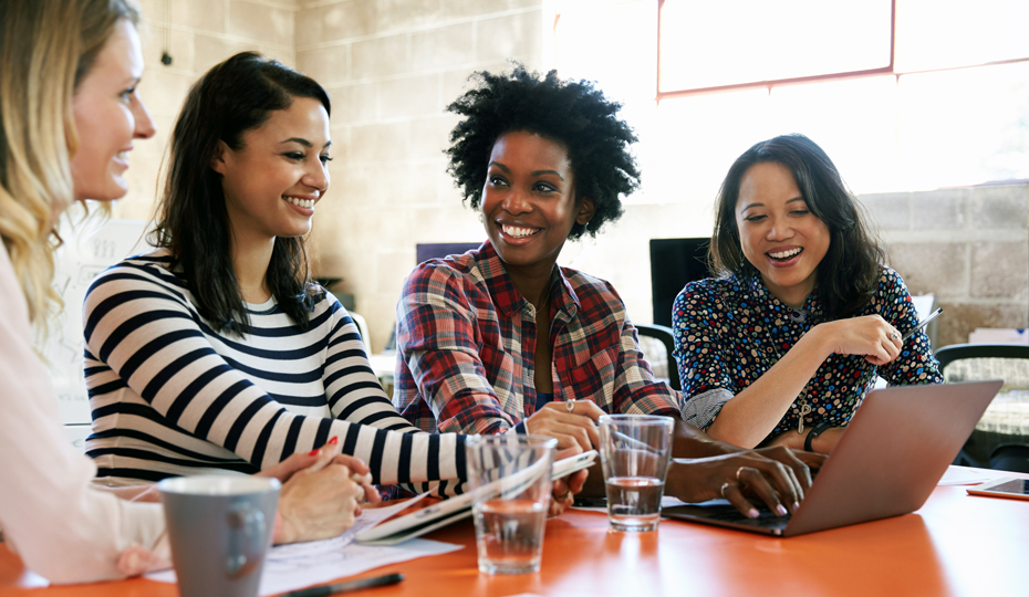 A group of women sitting around a table discussing notes and working on a laptop