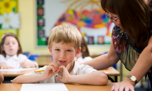 Young child sat at a desk with a teacher looking on