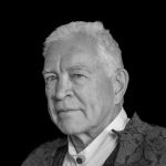 Older white man with short white hair staring at the camera in a white shirt and a grey jacket.