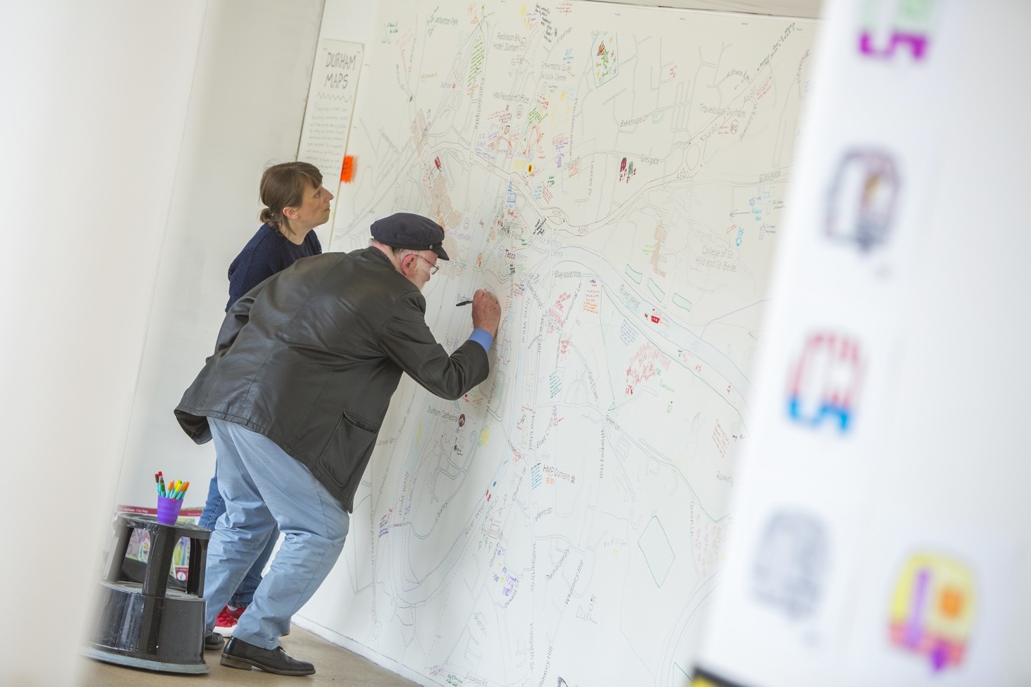 A man and a woman draw onto a large monochrome hand-drawn map, attached to a wall, both are crouched while the man draws onto the map.