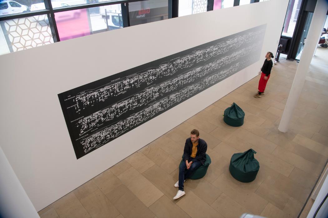 Exhibition image taken from above, a large, long and rectangular artwork appears on a white wall. The artwork consists of white musical notes on a black background. In front of the artwork, one person stands, a second person is sitting on a bean bag facing the front of the gallery.