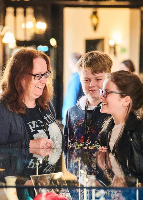 An adult and two children stand facing each other and laughing, in front of them is the top of a glass display case, to one side a historic tapestry hangs on the wall.