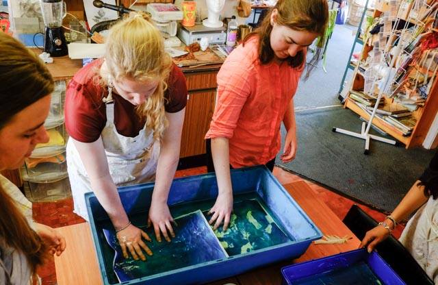Three women students stand around a table, two of them have their hands in blue plastic tubs filled with water, rinsing off printed artworks.
