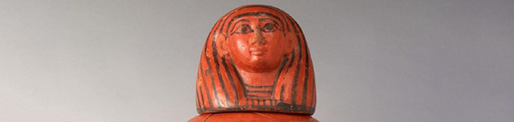 Close up view of the lid and upper section of a canopic jar featuring the human headed god Imsety, made of orange clay with details picked out in a black glaze.