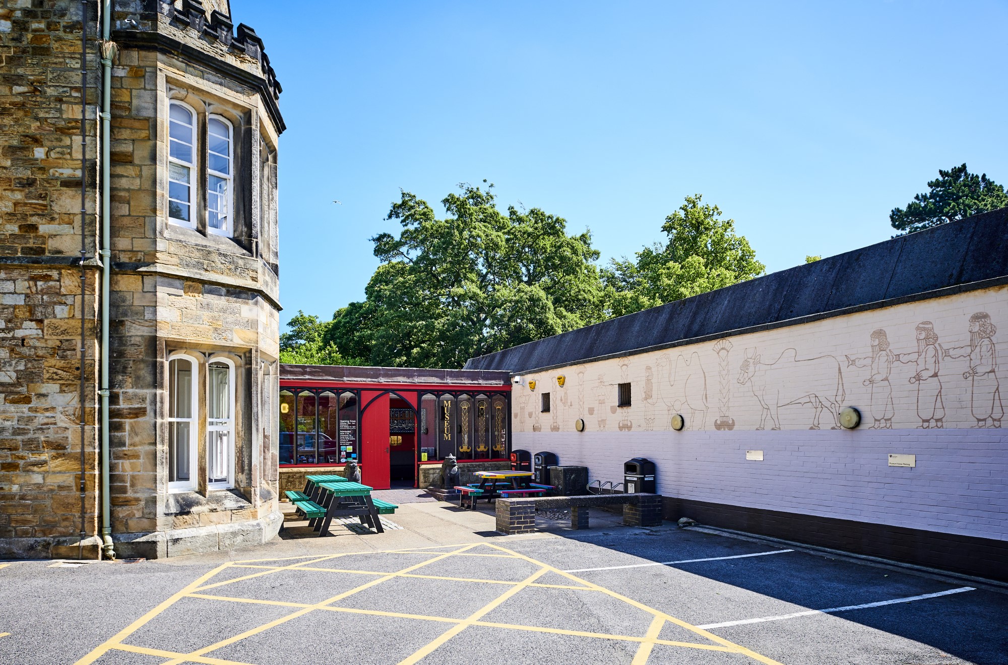 A line of parking bays leading to a courtyard area with picnic tables. Behind this is the entrance to a building with a shallow ramp and double red wooden doors.